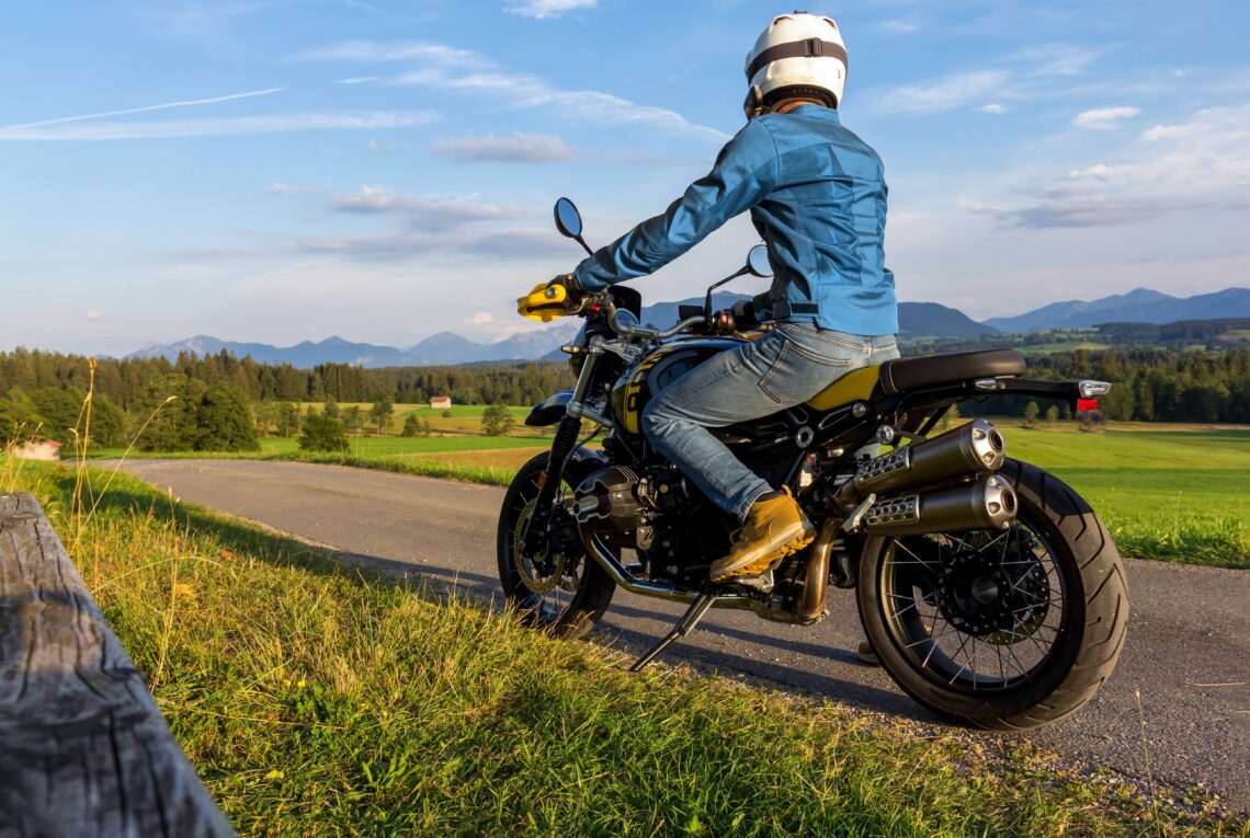 Ein Motorradfahrer auf einem 125er Motorrad, gekleidet in eine blaue Jacke und mit Helm, fährt auf einer ländlichen Straße, umgeben von grünen Feldern und einer malerischen Bergkulisse im Hintergrund. Die goldene Abendsonne taucht die Szene in warmes Licht und verstärkt das Gefühl von Freiheit und Abenteuer