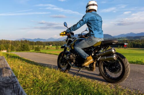 Ein Motorradfahrer auf einem 125er Motorrad, gekleidet in eine blaue Jacke und mit Helm, fährt auf einer ländlichen Straße, umgeben von grünen Feldern und einer malerischen Bergkulisse im Hintergrund. Die goldene Abendsonne taucht die Szene in warmes Licht und verstärkt das Gefühl von Freiheit und Abenteuer